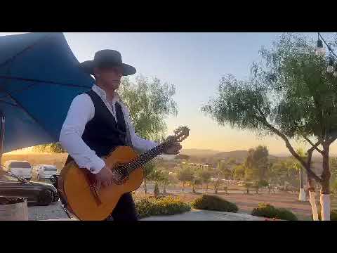 David Maldonado in a black hat and vest playing Spanish guitar outdoors at sunset on a terrace overlooking hills