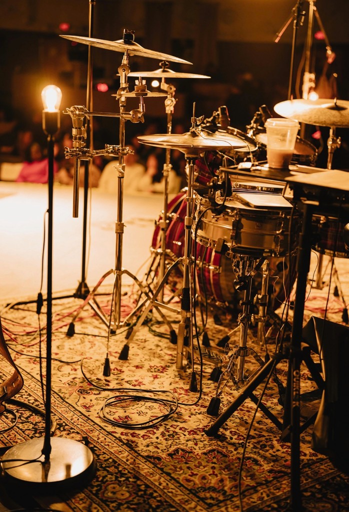 Drum kit on a patterned rug under warm stage lights with audience softly in the background