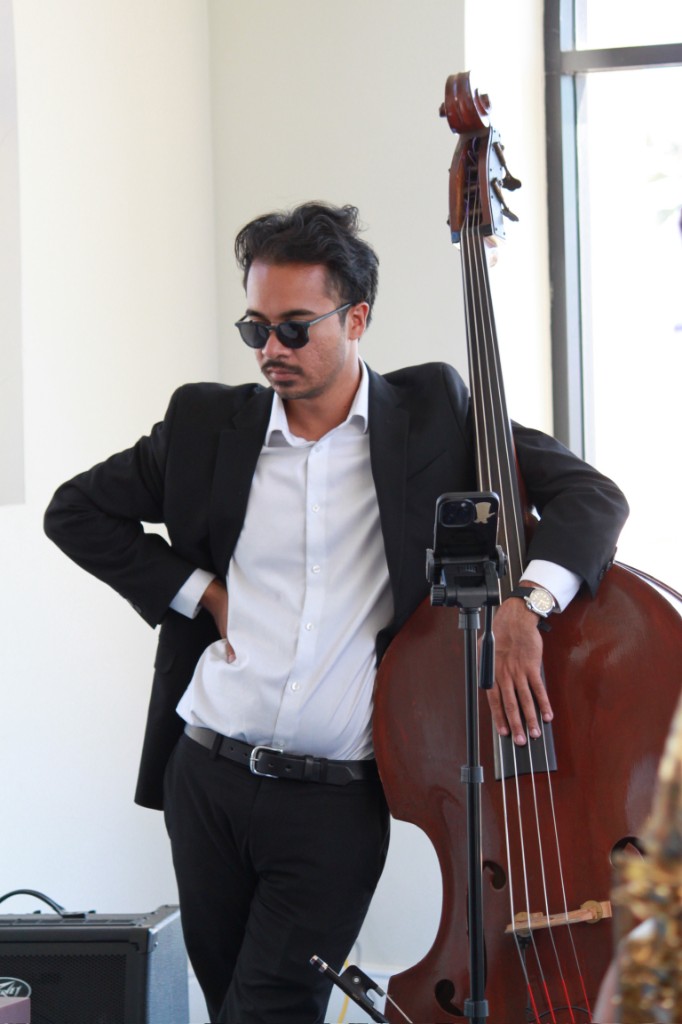 Bassist in a black suit and sunglasses leaning on an upright bass beside a mic stand and amp in a bright indoor room