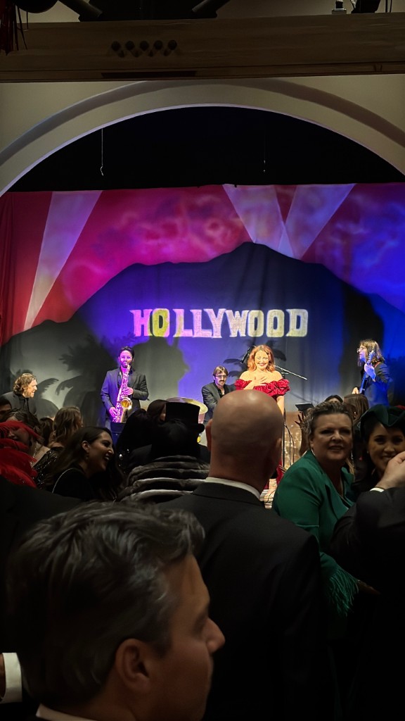 Stephanie Salahz in a red gown on stage before a lit Hollywood backdrop, full band and a formal audience in the foreground at a gala