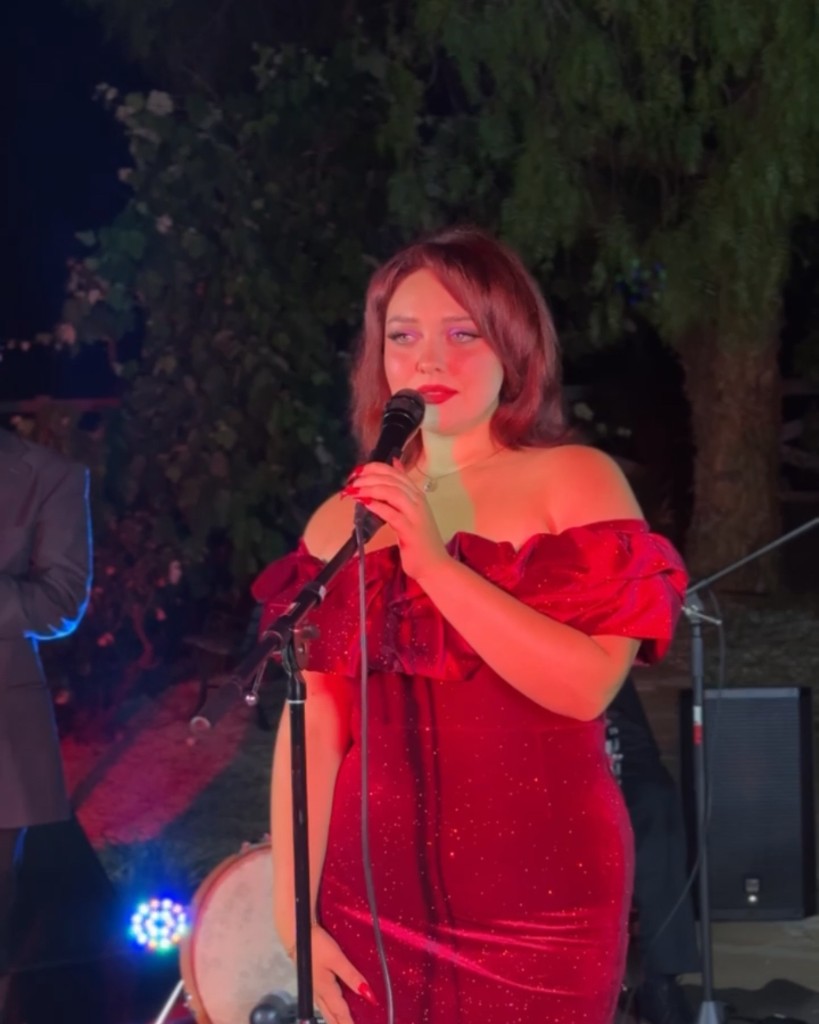 Stephanie Salahz in a burgundy off-the-shoulder dress singing into a microphone on an outdoor stage at night