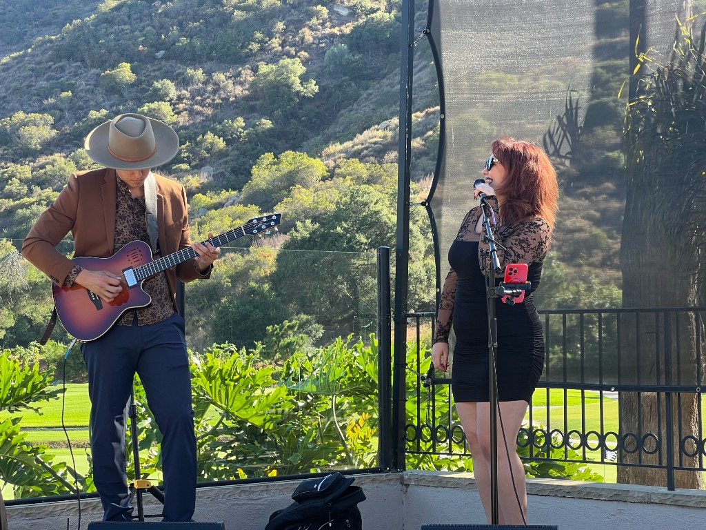 Stephanie Salahz singing with a guitarist on a sunlit stone terrace, green hills behind them
