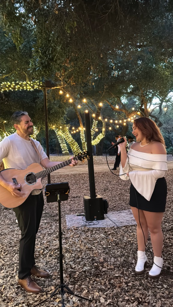 Stephanie Salahz singing outdoors at dusk beside an acoustic guitarist, fairy lights in the trees and fallen leaves underfoot