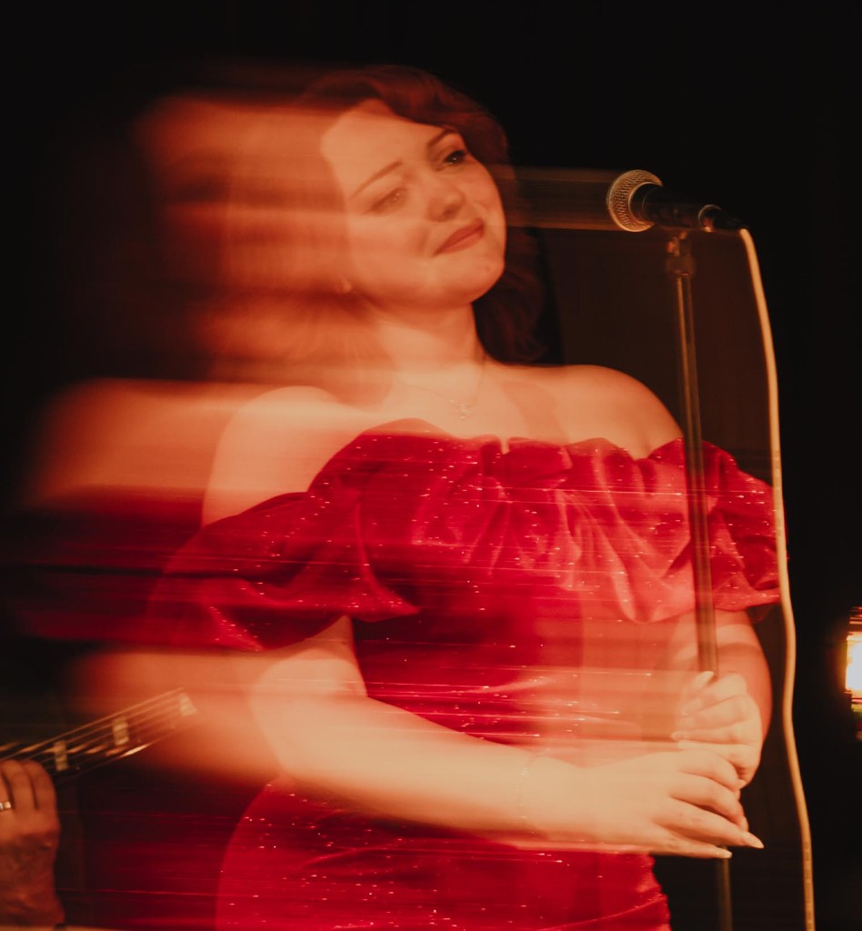 Stephanie Salahz in a sparkly red off-the-shoulder gown singing into a microphone, captured with horizontal motion blur on a warm-lit stage