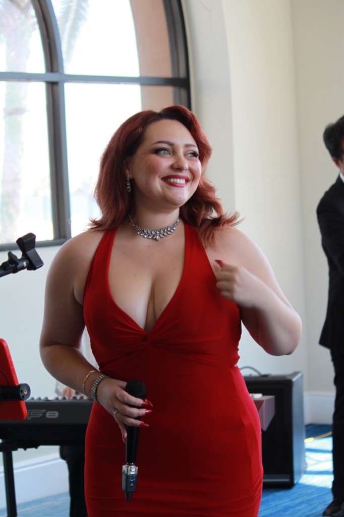 Stephanie Salahz smiling in a red halter gown with a handheld microphone, bright arched window and palm trees behind her